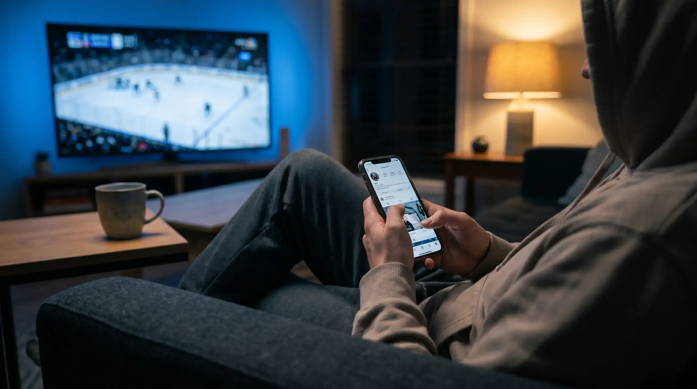 Person holding a smartphone displaying a sports betting interface while sitting on a sofa in the evening